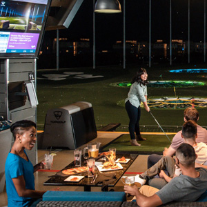 Eine Gruppe von Menschen sitzt an einem Tisch w�hrend im Hintergrund eine Frau am davor liegenden Au�enfeld Golf spielt